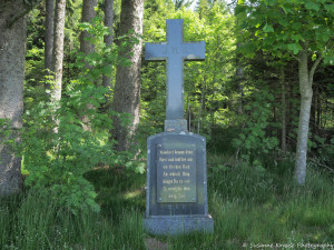 Das Foto zeigt ein Kreuz auf einer Steintafel. Auf der Steintafel steht folgender Spruch: Wanderer hemme deine Hast und halt bei mir ein bischen Rast. An irdisch' Ding hängst du zu viel Zu wenig für dein ewig Ziel.